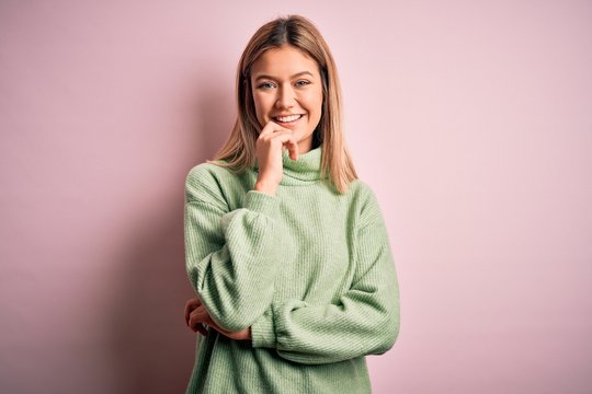 Young beautiful blonde woman wearing winter wool sweater over pink isolated background looking confident at the camera with smile with crossed arms and hand raised on chin. Thinking positive.