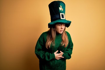 Young beautiful brunette woman wearing green hat on st patricks day celebration with hand on stomach because nausea, painful disease feeling unwell. Ache concept.