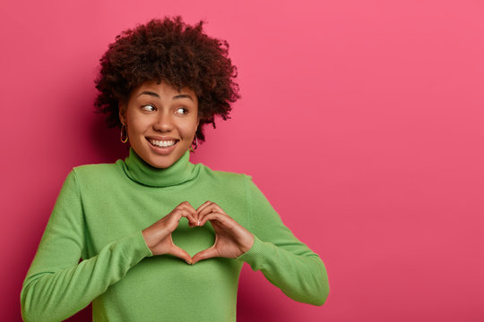 Portrait of good looking cheerful woman sends love, makes heart gesture, looks right with toothy smile, stands against pink wall with copy space for your advertising content, has romantic mood