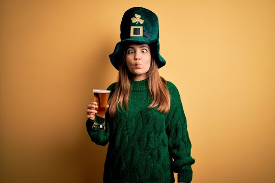 Young beautiful woman wearing green hat drinking glass of beer on saint patricks day making fish face with lips, crazy and comical gesture. Funny expression.