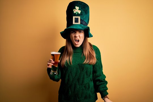Young beautiful woman wearing green hat drinking glass of beer on saint patricks day angry and mad screaming frustrated and furious, shouting with anger. Rage and aggressive concept.