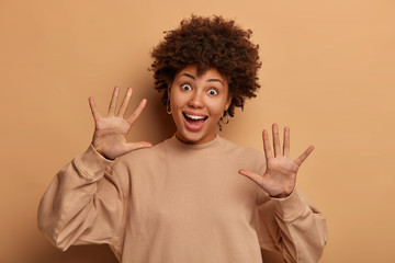 Funny playful young woman with curly hairstyle, pulls palms towards camera, laughs happily, has joyous expression, wears brown sweatshirt, sends positive vibes, stands against beige studio wall