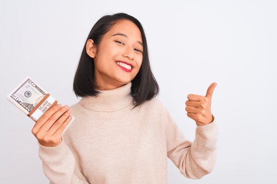 Young beautiful chinese woman holding dollars standing over isolated white background happy with big smile doing ok sign, thumb up with fingers, excellent sign