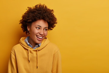 Fototapeta na wymiar Indoor shot of Afro American woman with natural curly hair, looks aside and smiles, remembers something pleasant, wears sweatshirt, isolated over yellow wall, copy space for your advertising