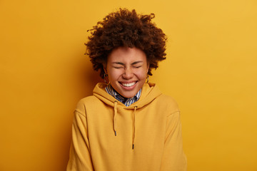 Fototapeta na wymiar Portrait of overjoyed Afro American female teenager closes eyes, smiles broadly, laughs at something positive, wears yellow hoodie, gets pleasure to communicate with comic person. Emotions concept