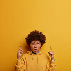 Wow, look there! Impressed shocked woman with Afro hair points fore fingers above, shows blank...