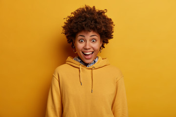 Fototapeta na wymiar Positive hipster girl has happy unexpected reaction on received gift, smiles toothily, wears yellow hoodie, stands indoor, has widely opened eyes, rejoices awesome news, isolated over vibrant wall