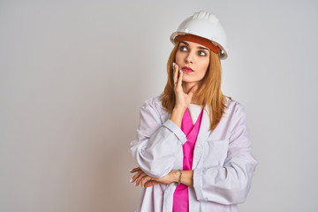 Redhead caucasian woman engineer wearing safety helmet over isolated background with hand on chin thinking about question, pensive expression. Smiling with thoughtful face. Doubt concept.