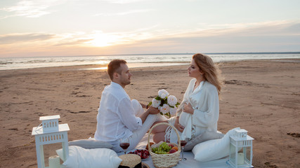Warm sunset. A man and pregnant woman had picnic on the sand. A man holds a glass with a drink, a woman - flowers.
