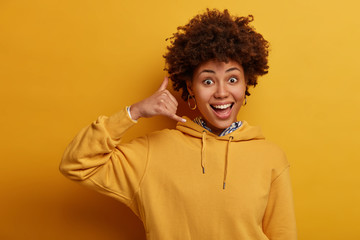 Fototapeta na wymiar Funny glad Afro American female does phone gesture, keeps fingers like talking on smartphone, smiles broadly, wears sweatshirt, poses over vivid yellow wall. Communnicating concept. Call me back