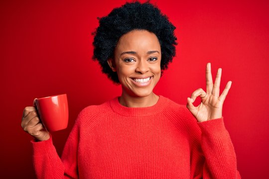 African American afro woman with curly hair drinking cup of coffee over red background doing ok sign with fingers, excellent symbol