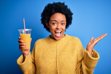 Young African American afro woman with curly hair drinking healthy orange juice very happy and...