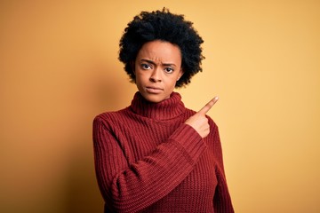 Young beautiful African American afro woman with curly hair wearing casual turtleneck sweater Pointing with hand finger to the side showing advertisement, serious and calm face