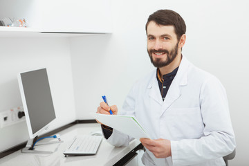 Happy mature male doctor smiling to the camera, sitting at his office. CHeerful practitioner doing paperwork at his clinic