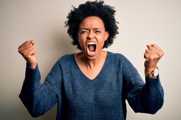 Young beautiful African American afro woman with curly hair wearing casual sweater angry and mad raising fists frustrated and furious while shouting with anger. Rage and aggressive concept.