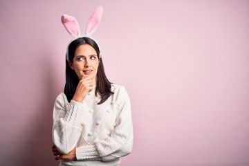 Young caucasian woman wearing cute easter rabbit ears over pink isolated background with hand on chin thinking about question, pensive expression. Smiling with thoughtful face. Doubt concept.