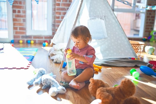 Adorable toddler holding feeding bottle around lots of toys at kindergarten