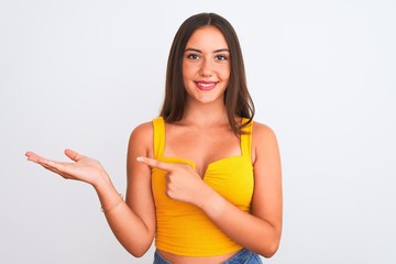 Young beautiful girl wearing yellow casual t-shirt standing over isolated white background amazed and smiling to the camera while presenting with hand and pointing with finger.