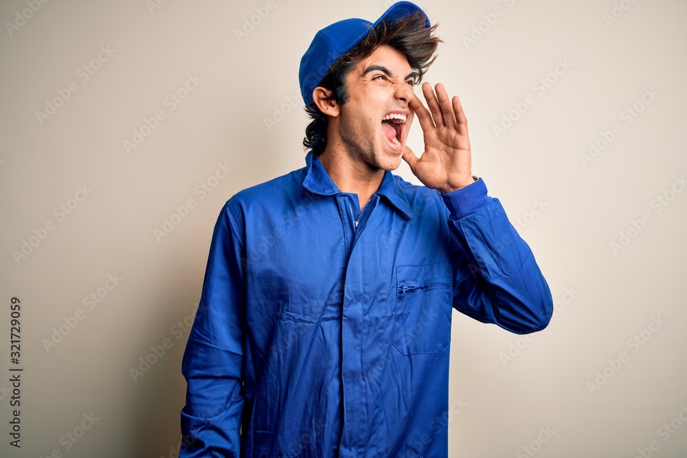 Wall mural Young mechanic man wearing blue cap and uniform standing over isolated white background shouting and screaming loud to side with hand on mouth. Communication concept.