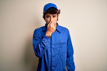 Young mechanic man wearing blue cap and uniform standing over isolated white background smelling something stinky and disgusting, intolerable smell, holding breath with fingers on nose. Bad smell