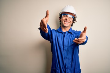 Young constructor man wearing uniform and security helmet over isolated white background looking at the camera smiling with open arms for hug. Cheerful expression embracing happiness.