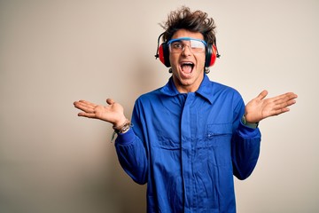 Young constructor man wearing uniform and earmuffs over isolated white background celebrating crazy and amazed for success with arms raised and open eyes screaming excited. Winner concept