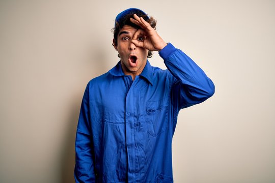 Young mechanic man wearing blue cap and uniform standing over isolated white background doing ok gesture shocked with surprised face, eye looking through fingers. Unbelieving expression.