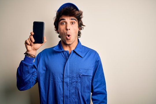 Young mechanic man wearing uniform holding smartphone over isolated white background scared in shock with a surprise face, afraid and excited with fear expression