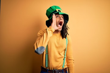 Young african american man wearing green hat celebrating saint patricks day shouting and screaming loud to side with hand on mouth. Communication concept.