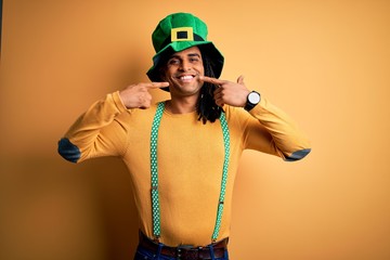 Young african american man wearing green hat celebrating saint patricks day smiling cheerful showing and pointing with fingers teeth and mouth. Dental health concept.