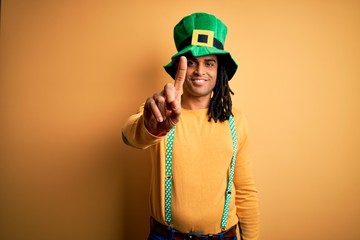 Young african american man wearing green hat celebrating saint patricks day showing and pointing up with finger number one while smiling confident and happy.
