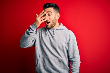 Young handsome sportsman wearing sweatshirt standing over isolated red background peeking in shock covering face and eyes with hand, looking through fingers with embarrassed expression.