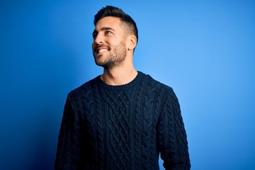 Obraz premium Young handsome man wearing casual sweater standing over isolated blue background looking away to side with smile on face, natural expression. Laughing confident.