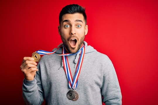 Young handsome succesful man winning medals standing over isolated red background scared in shock with a surprise face, afraid and excited with fear expression