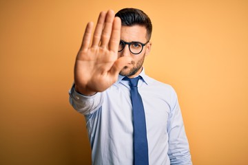 Young handsome businessman wearing tie and glasses standing over yellow background doing stop sing with palm of the hand. Warning expression with negative and serious gesture on the face.