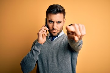 Young handsome man having conversation talking on the smartphone over yellow background pointing with finger to the camera and to you, hand sign, positive and confident gesture from the front