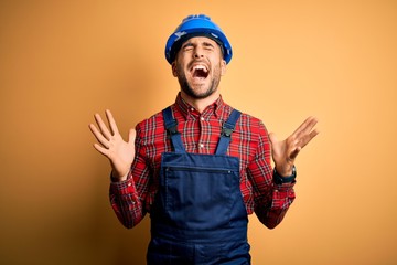 Fototapeta premium Young builder man wearing construction uniform and safety helmet over yellow background celebrating mad and crazy for success with arms raised and closed eyes screaming excited. Winner concept