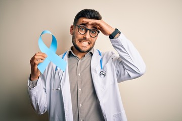 Young professional doctor man holding colon awareness blue ribbon over isolated background stressed...