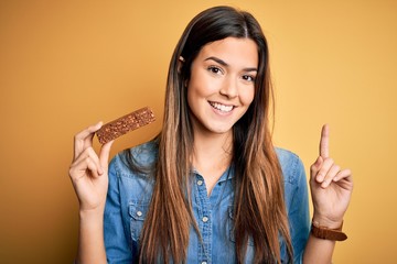 Young beautiful girl holding healthy protein bar standing over isolated yellow background surprised...