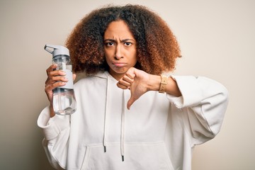 Young african american sportswoman with afro hair holding bottle of water with angry face, negative...