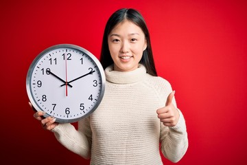 Young asian woman holding countdown big clock over red isolated background happy with big smile doing ok sign, thumb up with fingers, excellent sign