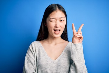 Young beautiful asian woman wearing casual sweater standing over blue isolated background smiling with happy face winking at the camera doing victory sign with fingers. Number two.