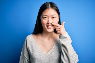 Young beautiful asian woman wearing casual sweater standing over blue isolated background Pointing with hand finger to face and nose, smiling cheerful. Beauty concept