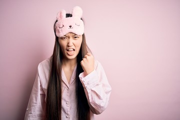 Young asian woman wearing pajama and sleep mask over pink isolated background angry and mad raising...