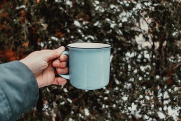Hand holding blue coffee cup in front of snow covered tree