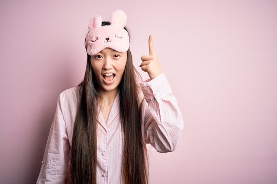 Young Asian Woman Wearing Pajama And Sleep Mask Over Pink Isolated Background Pointing Finger Up With Successful Idea. Exited And Happy. Number One.