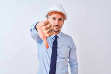Young business man wearing contractor safety helmet over isolated background looking unhappy and angry showing rejection and negative with thumbs down gesture. Bad expression.