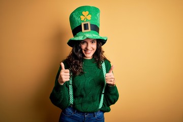 Beautiful curly hair woman wearing green hat with clover celebrating saint patricks day success sign doing positive gesture with hand, thumbs up smiling and happy. Cheerful expression and winner