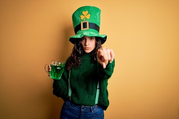 Beautiful curly hair woman wearing hat drinking jar of green beverage on saint patricks day pointing with finger to the camera and to you, hand sign, positive and confident gesture from the front