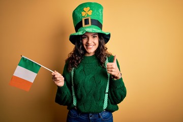 Beautiful patriotic curly hair woman wearing hat holding irish flag celebrating saint patricks day happy with big smile doing ok sign, thumb up with fingers, excellent sign